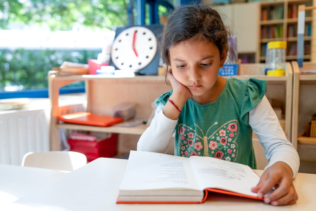 Kind leest boek aan tafel op school. Op de achtergrond, een klok.